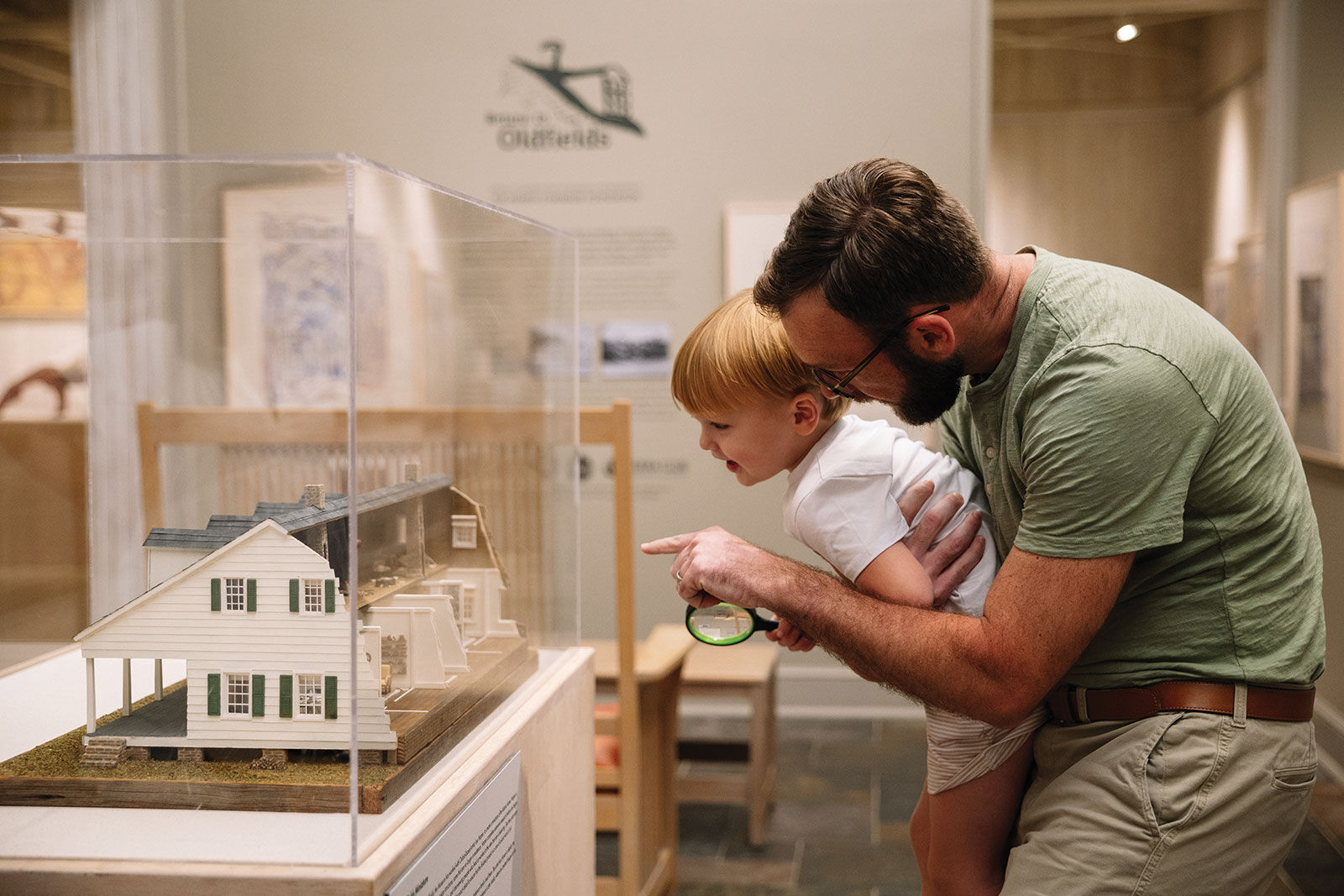 A man holds his son in front of a glass case in a museum, pointing to the model home inside